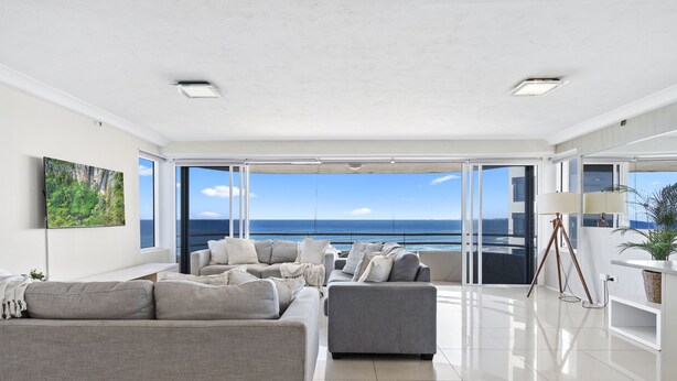 A modern living room with a large sofa, a smaller armchair, and a side table. The room has large windows offering a view of the ocean. - Spacious 2 Bedroom Ocean View Apartment - Q Stay (Surfers Paradise)