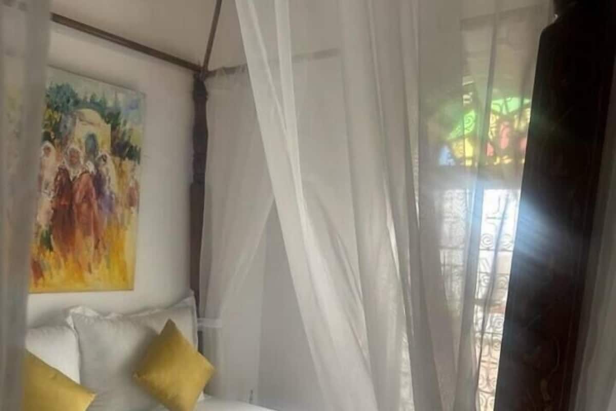 Un letto con biancheria bianca e cuscini gialli, un quadro al muro e una finestra con tende leggere.