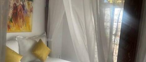 Ein Bett mit weißer Bettwäsche und gelben Kissen, ein Gemälde an der Wand und ein Fenster mit durchsichtigen Vorhängen.