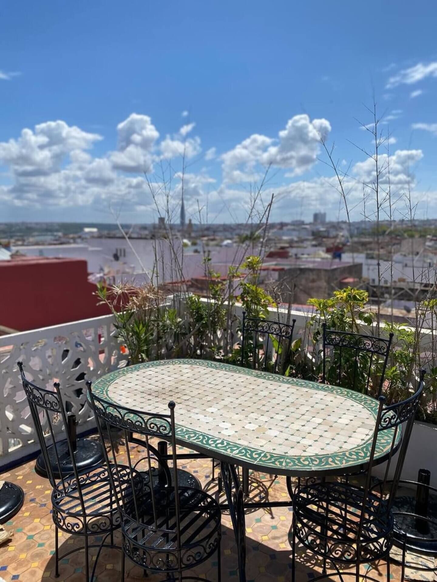 Ein runder Tisch mit einer grün-weißen Tischdecke, umgeben von schwarzen Metallstühlen, befindet sich auf einem Balkon mit Blick auf eine Stadtlandschaft.