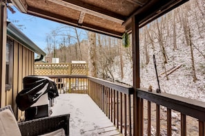 A covered outdoor deck with a grill, wicker furniture, and a view of a snowy forest.