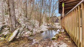 Ein Holzhaus mit überdachter Veranda, ein Bach fließt daneben, und ein bewaldetes Gebiet mit kahlen Bäumen.