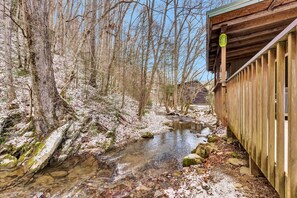 Ein Holzhaus mit überdachter Veranda, ein Bach fließt daneben, und ein bewaldetes Gebiet mit kahlen Bäumen.