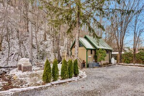 A rustic cabin with a green roof, surrounded by trees and a gravel driveway.