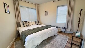 A bedroom with a bed, two windows with blinds, a bedside table, and a coat rack. - Entire apartment, Boston in 14min, Beach in 4min! (Revere)