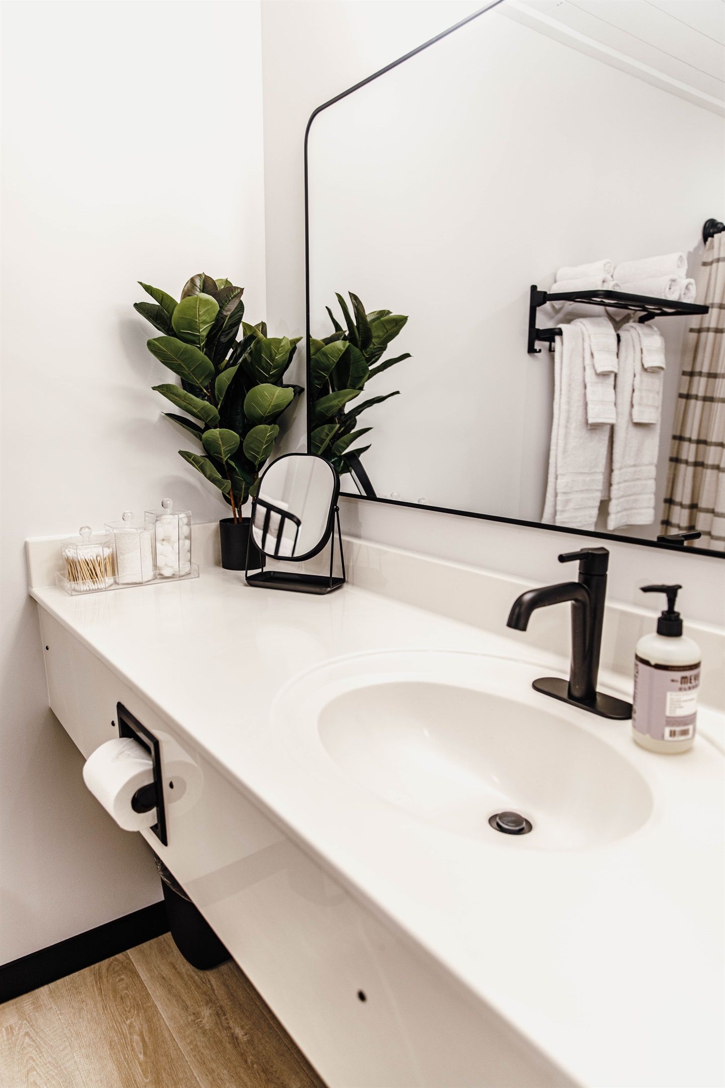 A modern bathroom with a white sink, black faucet, and a mirror above it. There are two potted plants on the counter and a towel rack with towels.