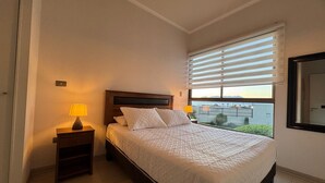 A bedroom with a bed, a nightstand with a lamp, a window with blinds, and a view of the outside. - Patagua (Antofagasta)