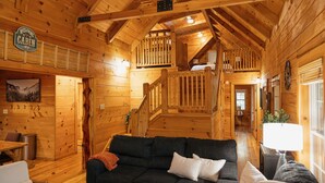 Interno di una baita con soffitto in legno, una scala, un divano e un cartello di benvenuto.