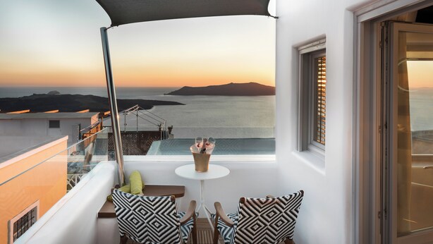 Ein Balkon mit Meerblick, zwei stilisierte Stühle, ein kleiner Tisch mit einem Glas und ein Fenster mit Jalousien.
