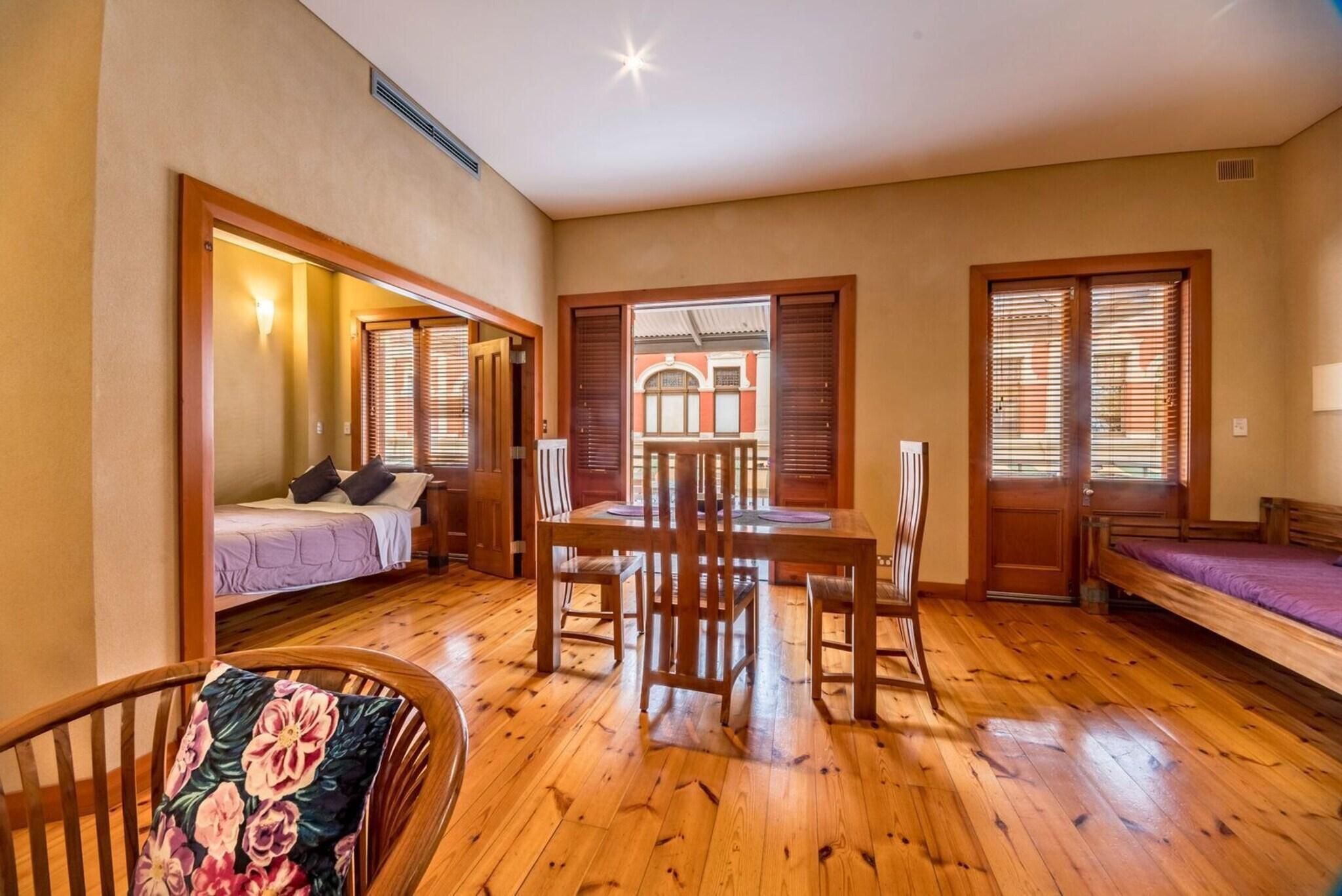 A room with a bed, a dining table with chairs, a wooden floor, and a view through a door.