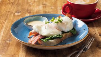 Un plato de desayuno con huevos Benedict, tocino y verduras, acompañado de una taza roja de café sobre una mesa de madera.