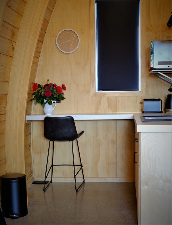 Une cuisine compacte avec des murs en bois, un plan de travail blanc, une chaise noire, un pichet blanc avec des fleurs, un tableau noir et un grille-pain.