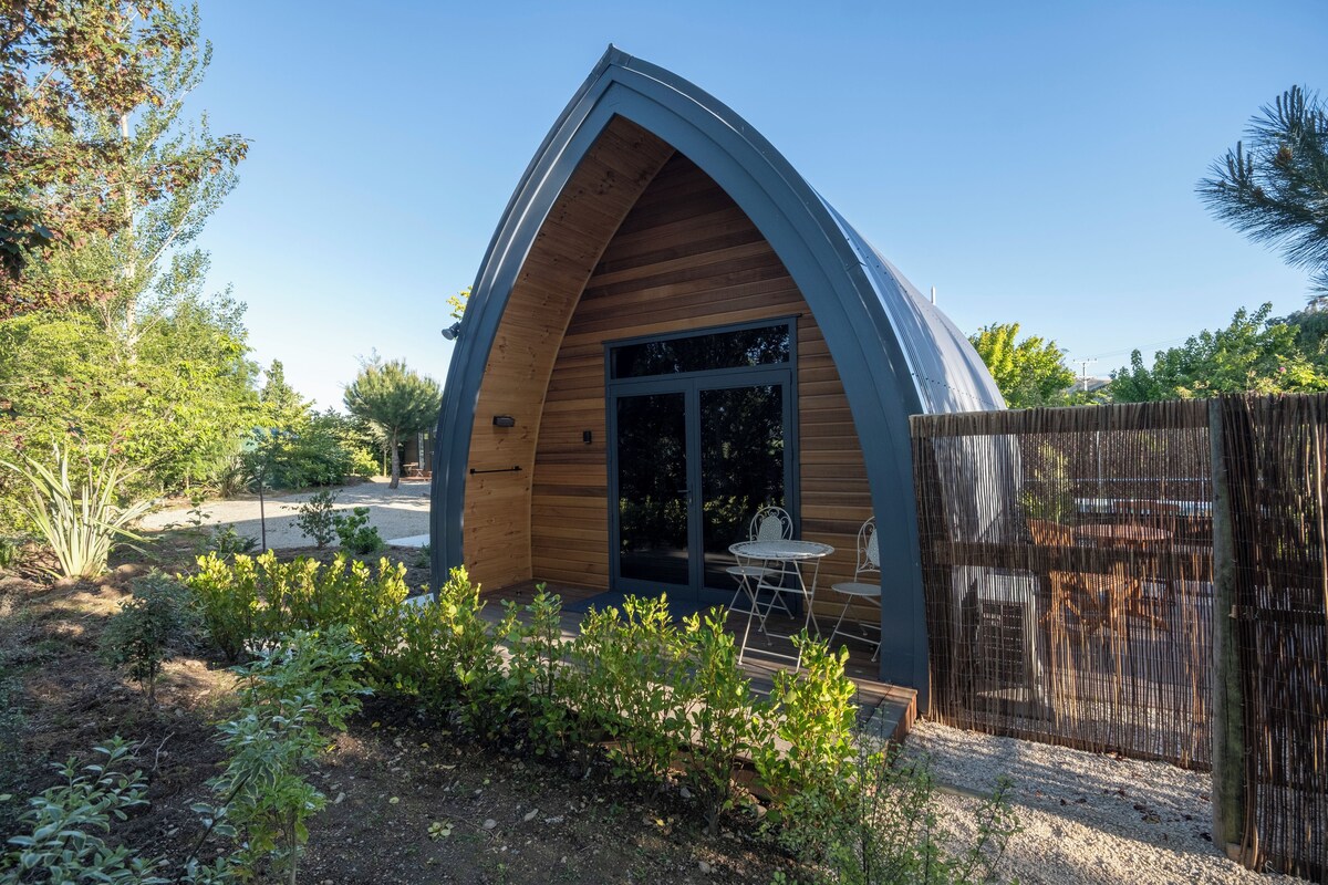 A modern, arched wooden cabin with a glass door and a small patio area with a table and chairs.