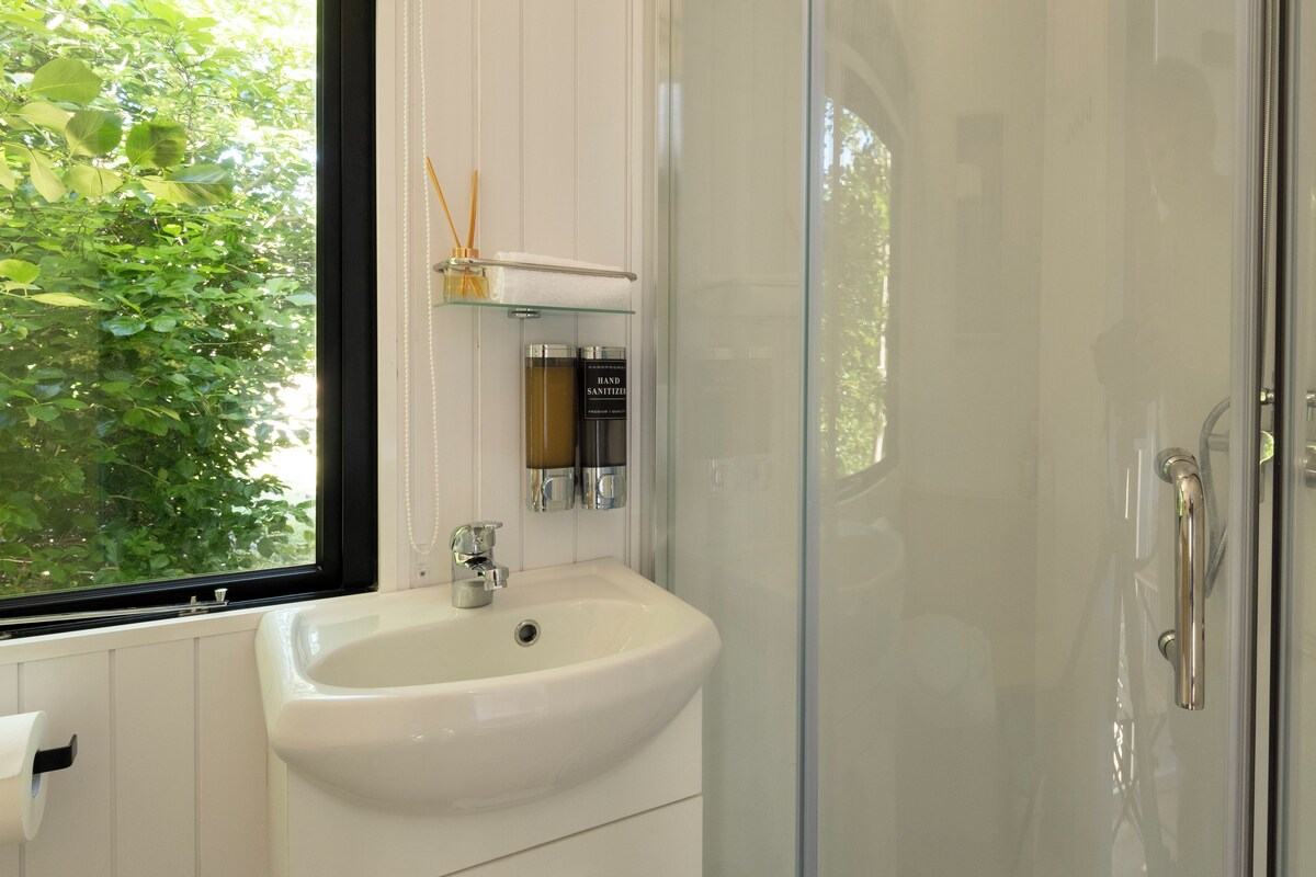 Un baño con lavamanos blanco, cabina de ducha de vidrio y ventana con vista a follaje.