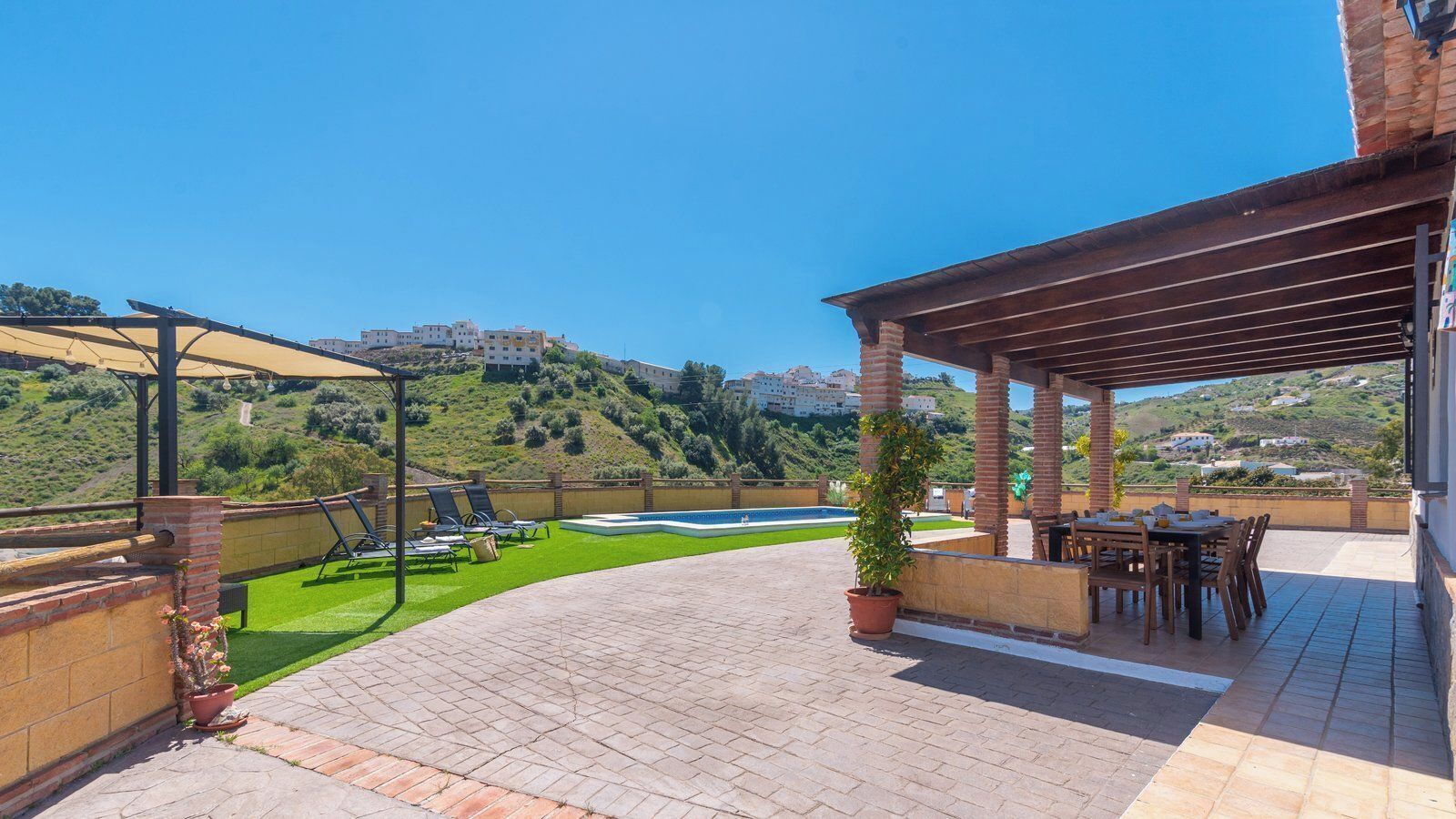 A covered outdoor patio with a pool, green lawn, and a view of a hillside with buildings.