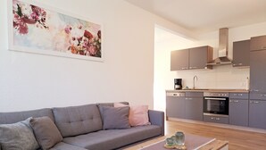 A modern living room with a grey sectional sofa, a coffee table with decorative items, and a kitchen with grey cabinets and a stainless steel oven. - Idyllic vacation home "Björn", Büsum (6km), terrace, parking lot, secluded location (Reinsbüttel)