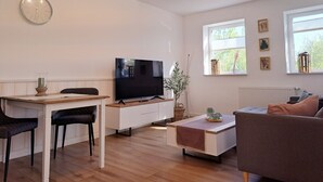 A modern living room with a flat-screen TV, wooden flooring, a dining table with chairs, a sofa, and a window with artwork. - Idyllic vacation home "Björn", Büsum (6km), terrace, parking lot, secluded location (Reinsbüttel)