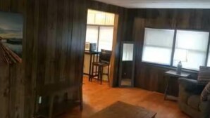Una sala de estar con paredes y mesa de centro de madera, y un sofá. Hay una ventana con persianas y una puerta que da a otra habitación.
