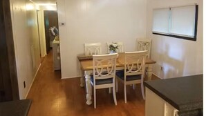 Comedor con mesa de madera y sillas blancas, una ventana con persianas, y una zona de cocina con mostrador.