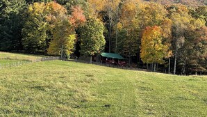 A grassy field with a wooden fence, a cabin with a green roof, and a forested hill in the background. - Cozy 2bedroom mountain cabin on 18 acres near the Appalachian Trail (Chilhowie)