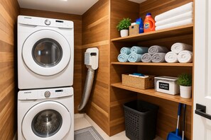 Une buanderie équipée de deux machines à laver superposées, d’un sèche-linge, d’une étagère avec des serviettes, du matériel de lessive et un balai.
