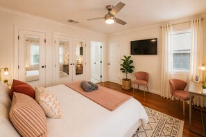 Une chambre d’hôtel équipée d’un lit, d’une télévision, d’un bureau avec une chaise, d’une plante et d’un ventilateur de plafond.