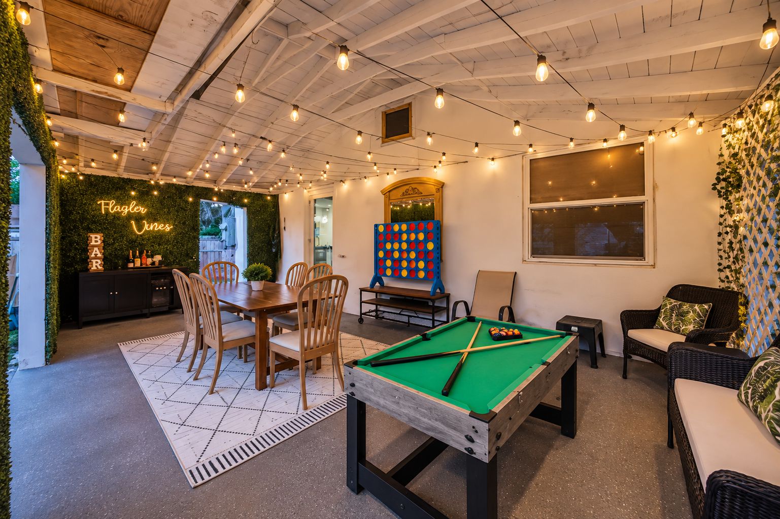 Una mesa de billar con superficie de fieltro verde, rodeada de sillas de madera y una mesa de comedor con sillas. Hay una zona de bar con botellas y vasos, y un letrero de neón que dice 'Flagler Vines'.