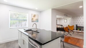 A modern kitchen with a black countertop, white cabinets, and a window with blinds. - 6Bed Near Beach - Grill, Pool Table, FirePit - Family & Event Open (West Palm Beach)