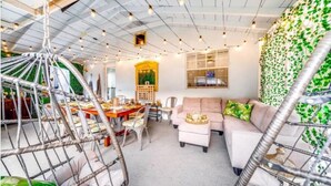 A modern living room with a high ceiling, a hanging chair, a sofa, and a dining area with chairs and a table. - 6Bed Near Beach - Grill, Pool Table, FirePit - Family & Event Open (West Palm Beach)