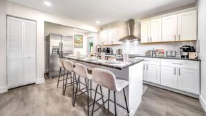 A modern kitchen with white cabinets, a central island with bar stools, stainless steel appliances, and a large window. - 6Bed Near Beach - Grill, Pool Table, FirePit - Family & Event Open (West Palm Beach)