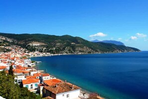 A coastal town with red-roofed buildings, a clear blue sea, and green hills in the background.
