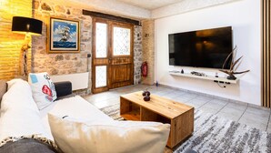 Un saló amb un sofà, taula de centre, televisió, paret de pedra, porta de fusta i una estanteria amb objectes decoratius.