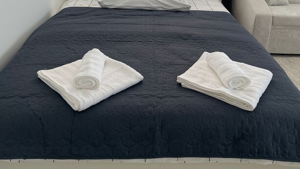 A neatly made bed with a dark quilt, two rolled towels, and a bedside table with a lamp.