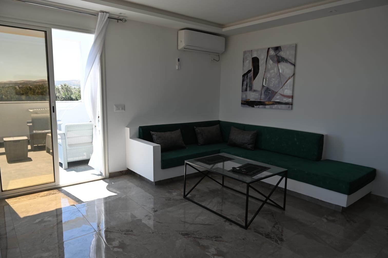 A modern living room with a green sofa, a glass coffee table, and a sliding glass door leading to an outdoor patio.