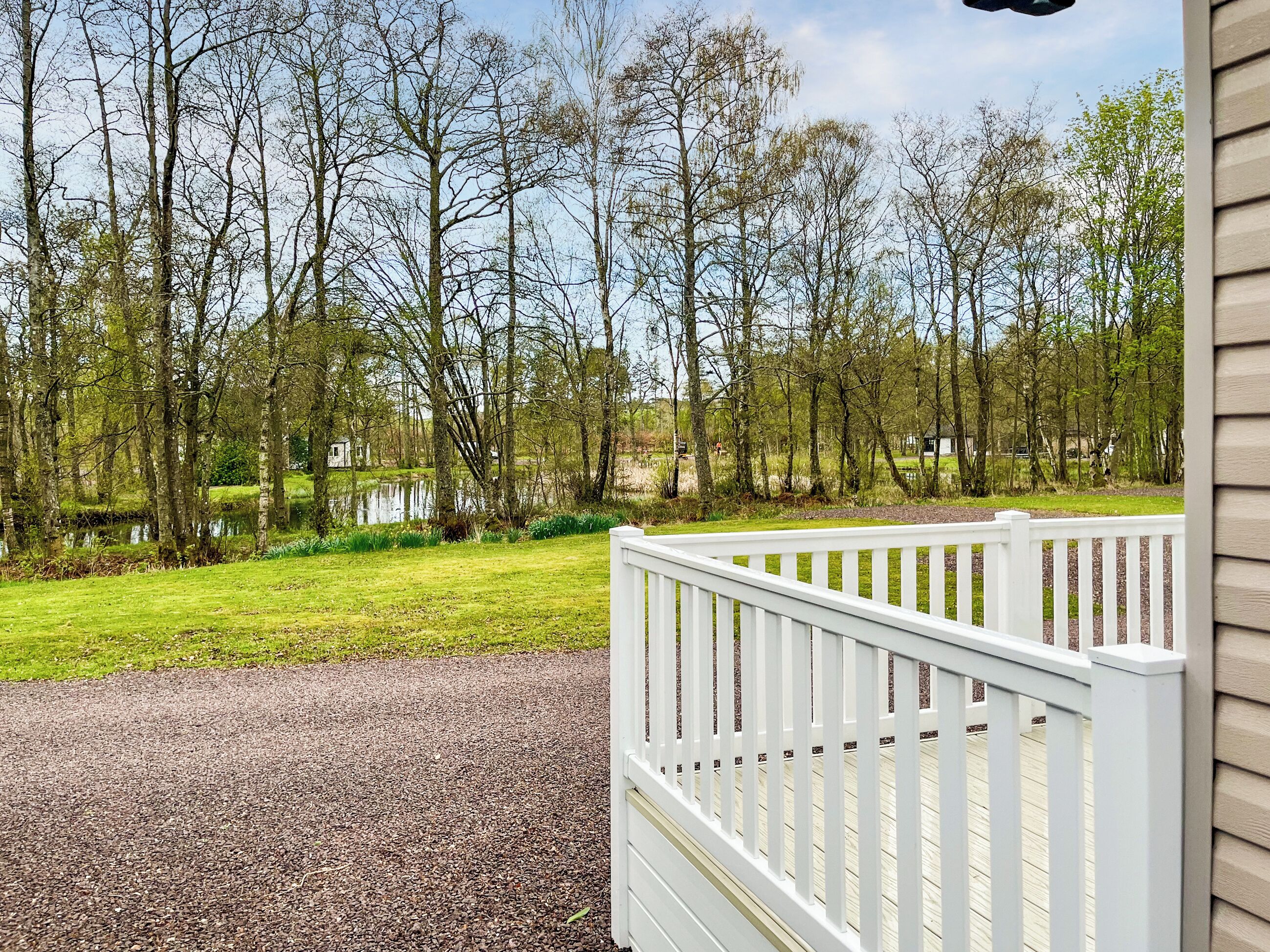 Blick von einer Veranda, mit einem Kiesweg, der zu einer Wiese und einem Gewässer führt, umgeben von Bäumen und einem weißen Zaun.