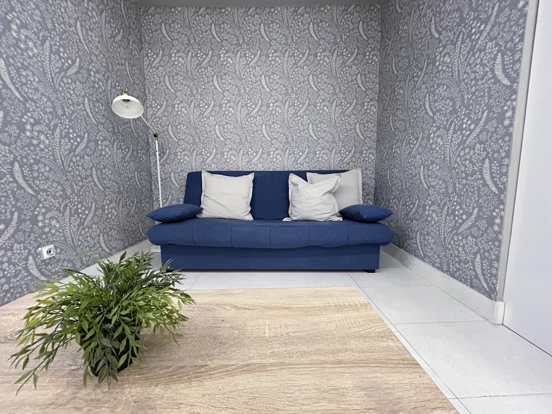 A blue sofa with white cushions against a patterned wallpaper, a floor lamp, and a green plant on a wooden floor.