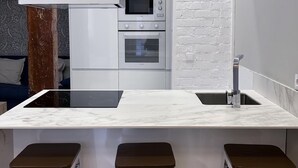 Una cocina moderna con encimera blanca, horno empotrado y fregadero. Hay tres taburetes blancos con asientos marrones.
