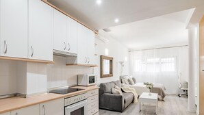 A modern kitchen with white cabinets, a built-in oven, and a microwave. There is a grey sofa and a small white table in the living area.