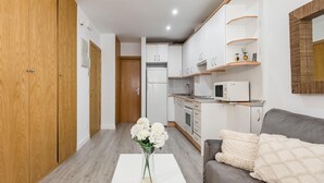 A modern kitchen with white cabinets, a microwave, and a built-in oven. There is a wooden wardrobe on the left and a grey sofa with a white cushion on the right.