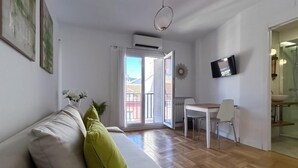 A modern living room with a sofa, a dining table with chairs, a TV mounted on the wall, and an air conditioning unit. - Sol Studio III - Apartment in the City Center with Balcony (Madrid)