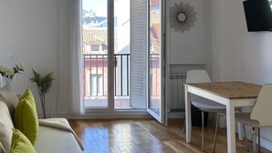 A modern living room with a wooden floor, a white sofa with green cushions, a dining table with chairs, and a TV mounted on the wall. - Sol Studio III - Apartment in the City Center with Balcony (Madrid)