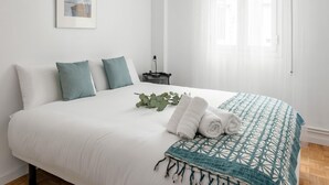 A neatly made bed with white linens and a teal patterned throw, a bedside table with a lamp, and a window with sheer curtains. - La Latina Life - 2-Bedroom Apartment with Balcony (Madrid)