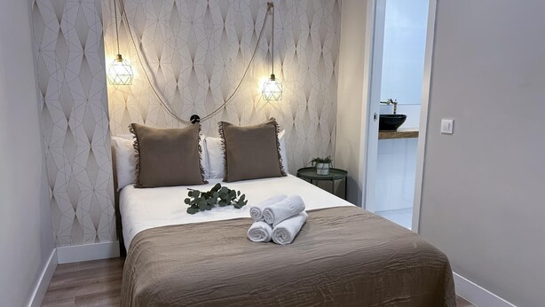A neatly made bed with a brown bedspread, two pillows, and a small plant on a bedside table. There is a white door leading to a bathroom with a sink and a mirror. - Madrid Experience - 3-Bedroom Apartment in the City Center (Madrid)
