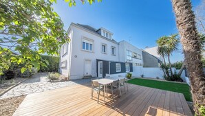 Une maison à deux étages avec une terrasse en bois, un petit jardin et une façade blanche.