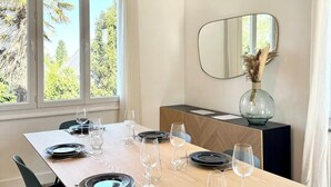 Un coin repas avec une longue table en bois dressée pour quatre personnes, des assiettes noires, des verres à vin transparents et un vase contenant des fleurs séchées, posé sur une console sombre.