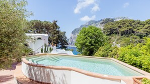 Ein Pool mit geschwungener Kante, umgeben von Grün und Blick auf eine gebirgige Küstenlinie.
