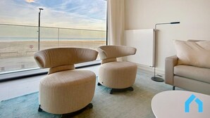 Une chambre d’hôtel moderne dotée d’une grande fenêtre donnant sur la plage. On y trouve deux fauteuils et un canapé.