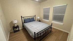 Ein Schlafzimmer mit einem Bett, zwei Nachttischen, zwei Lampen und zwei Fenstern mit Jalousien.