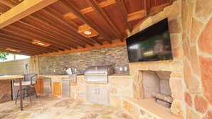 Une cuisine extérieure avec un mur en pierre, un barbecue, un téléviseur et un espace salon.