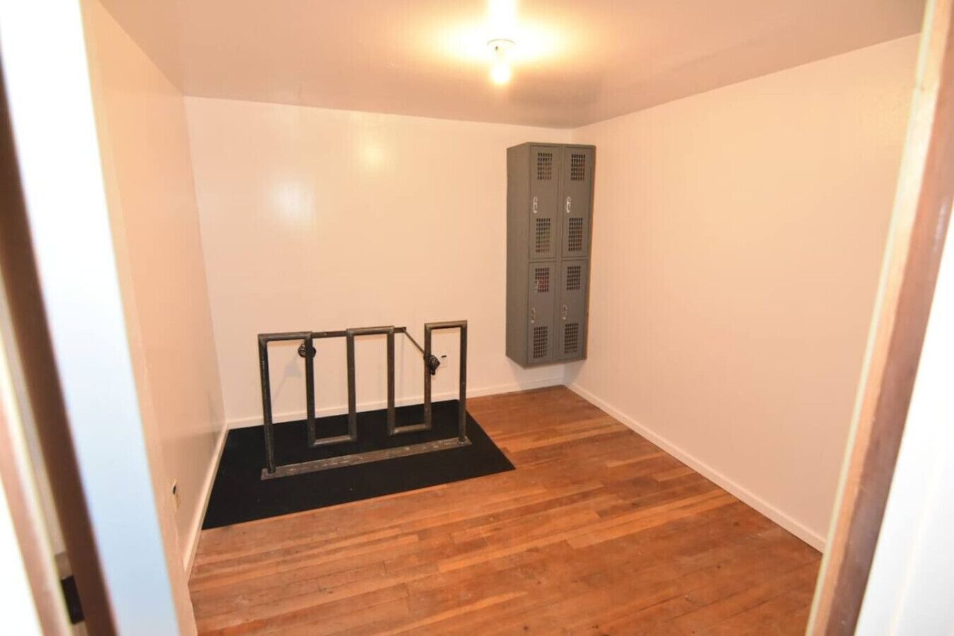 Une petite pièce vide, avec un plancher en bois, un tapis noir et un étagère en métal adossée au mur.